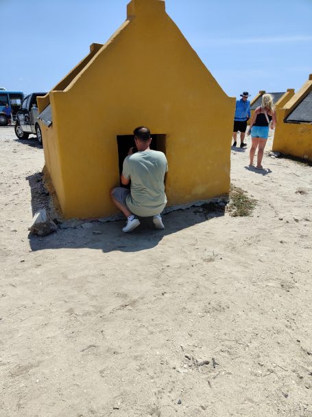 Een persoon leunt tegen een gele hutje op het strand, omringd door andere mensen.
