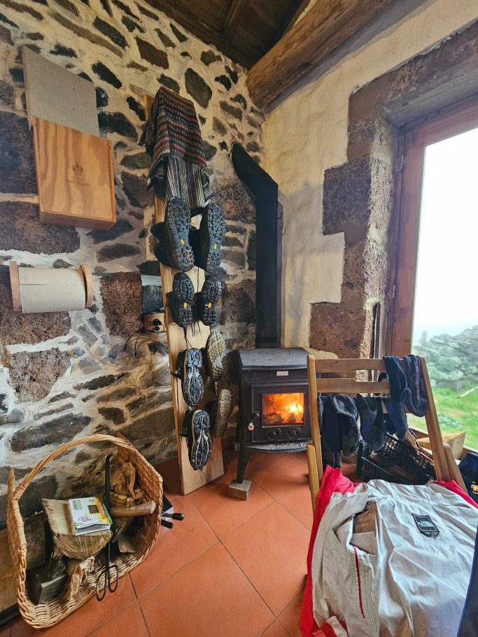 Houten stoel naast een open haard en een stenen muur, met een mand en kleding op de vloer.