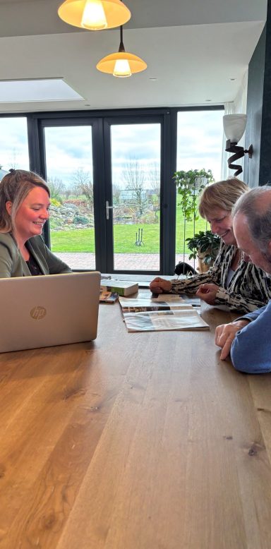 Drie mensen aan een houten tafel, met een laptop en documenten, in een lichte ruimte.