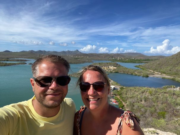 Zonnige selfie van een stel met water en bergen op de achtergrond.
