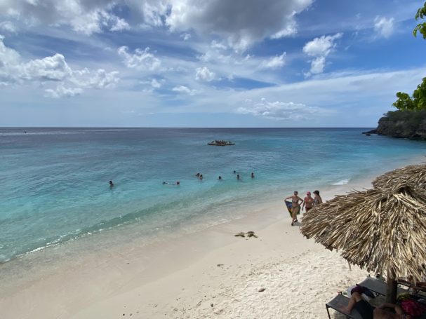 Rustig strand met helder blauw water, mensen zwemmen en palmbomen op de achtergrond.