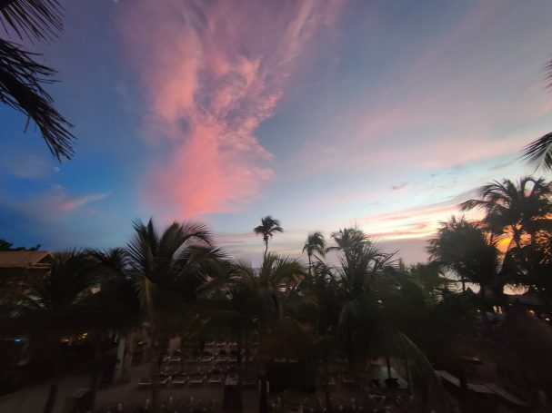 Zonsondergang met roze en paarse wolken boven palmbomen en een zee-uitzicht.