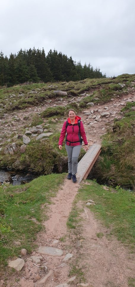 Vrouw in roze jas wandelt over een smalle houten brug in een groene omgeving.