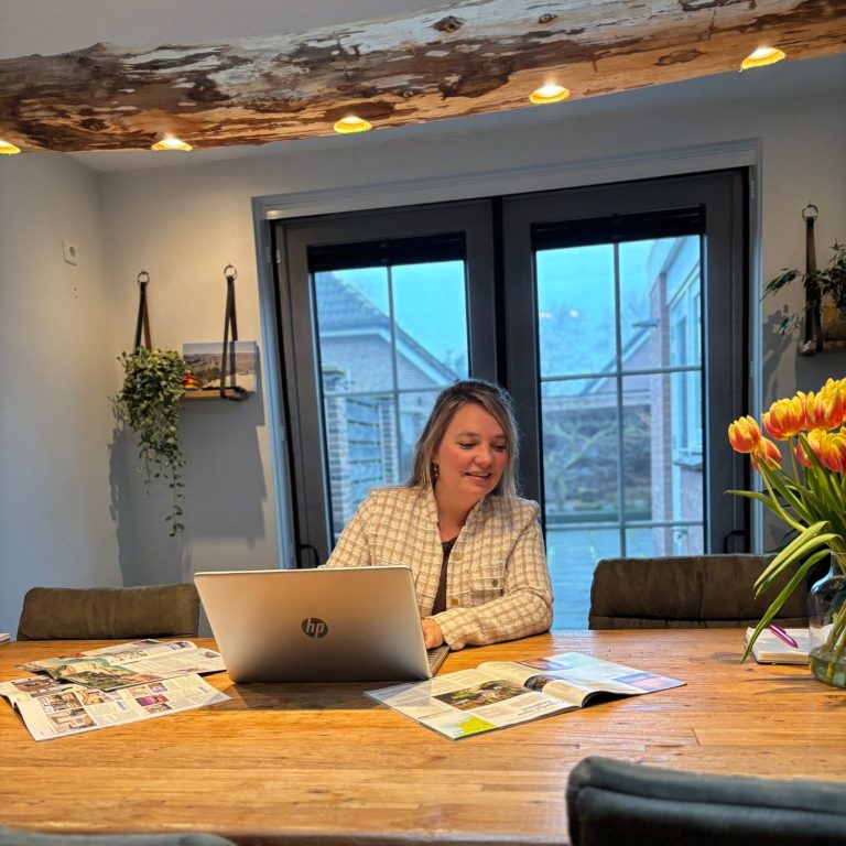 Vrouw in een lichte kamer, werkend aan een laptop met tijdschriften en bloemen op de tafel.