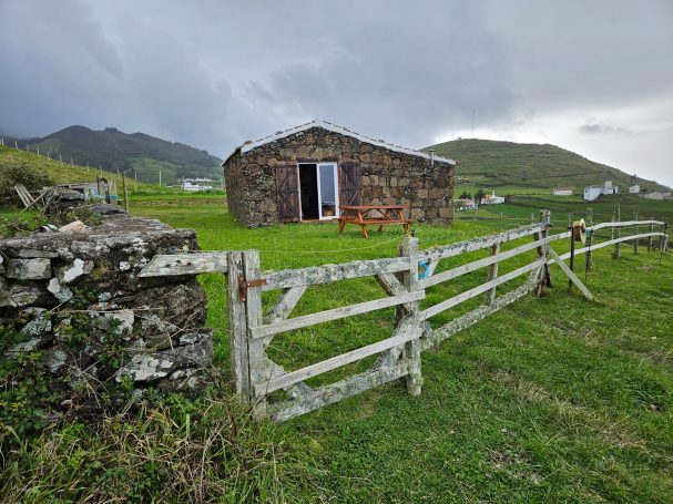 Een stenen huis met een houten hek, omgeven door groene velden en heuvels.