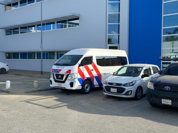 Witte van met rood-blauwe strepen, geparkeerd naast een kleinere auto. Moderne gebouwen op de achtergrond.
