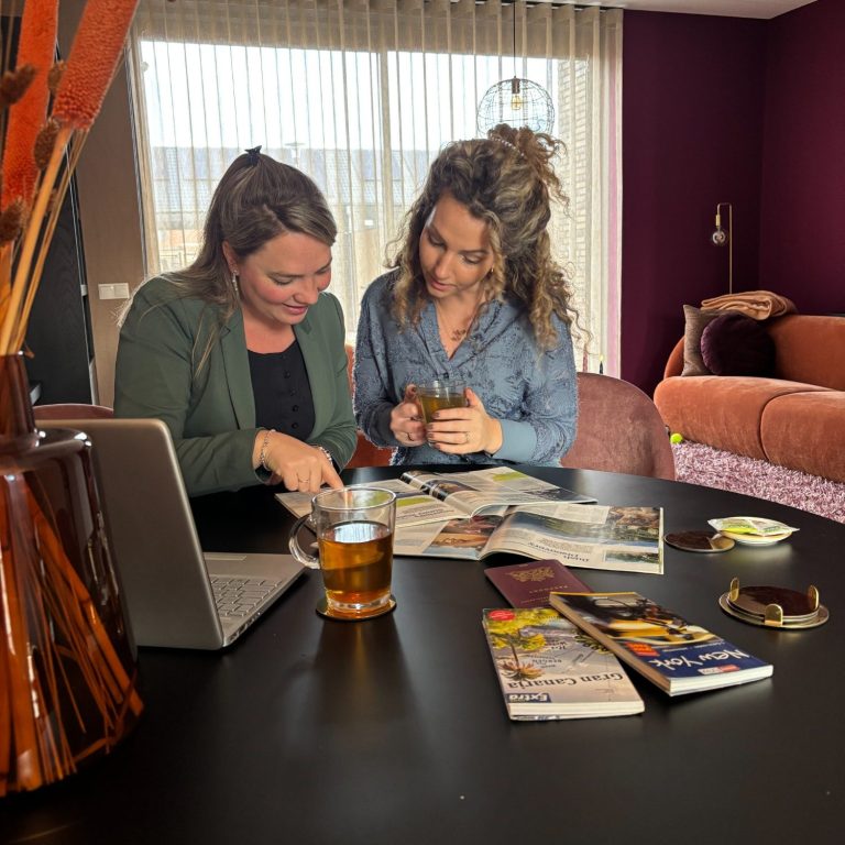 Twee vrouwen zitten aan een tafel met tijdschriften en een laptop, genietend van een drankje.