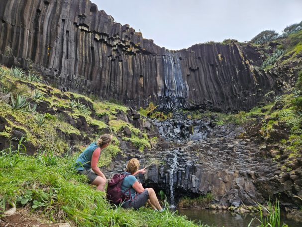 Twee personen zitten bij een waterval omringd door basaltformaties en groene vegetatie.