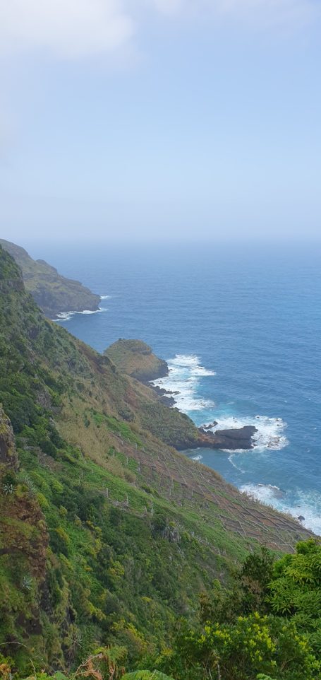 Kustlijn met golven die tegen de groene bergen slaan onder een blauwe lucht.