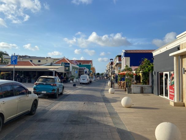 Drukke straat met winkels, witgekleurde gebouwen en enkele voertuigen onder een blauwe lucht.