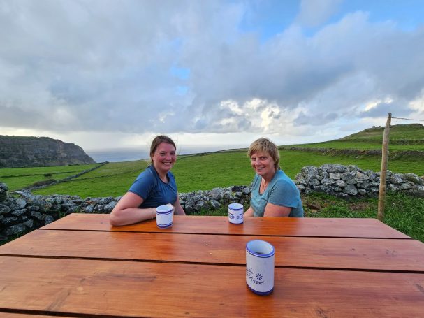 Twee vrouwen zitten aan een houten tafel met uitzicht op groene heuvels en een bewolkte lucht.